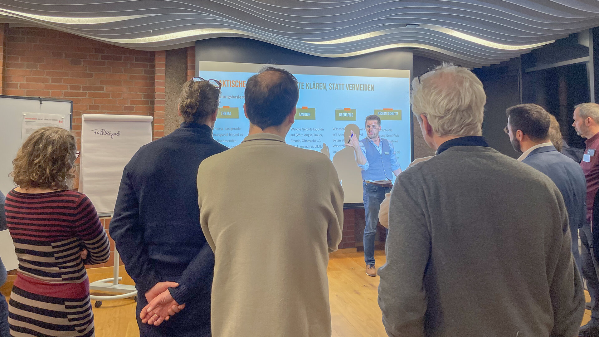 Eine Gruppe von Menschen steht in einem Raum und verfolgt aufmerksam, wie Chris Batke (Coach für Transformation) im Bauunternehmen Gundlach eine Präsentation hält, bei der Folien auf eine Leinwand projiziert werden. 